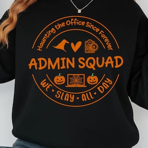 May include: Black sweatshirt with an orange graphic that reads "ADMIN SQUAD" inside a circular design with Halloween-themed icons. The design includes a witch hat, heart, camera, pumpkin, and a laptop with a spiderweb. The text "Haunting the Office Since Forever" and "We Slay All Day" are also included.
