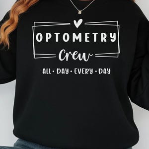 May include: Black crewneck sweatshirt with white text that reads "OPTOMETRY Crew ALL • DAY • EVERY • DAY" inside a rectangular box with a heart above it. The sweatshirt has long sleeves and a ribbed cuff.