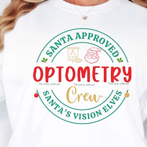 Puede incluir: Sudadera blanca con diseño festivo. El diseño incluye el texto "SANTA APPROVED OPTOMETRY CREW SANTA'S VISION ELVES" en rojo, verde y dorado. También se incluye un gráfico de Papá Noel y una tabla optométrica.