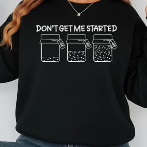 May include: Black sweatshirt with the text "DON'T GET ME STARTED" above three jars with contents. The jars have a tag hanging from them. The sweatshirt has long sleeves and a crew neck.
