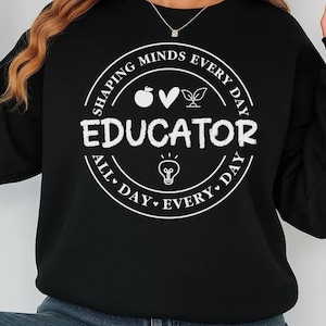 May include: Black sweatshirt with a white circular graphic that reads "EDUCATOR" in bold letters. The graphic includes the text "SHAPING MINDS EVERY DAY" and "ALL DAY EVERY DAY" around the circle, along with an apple, heart, leaves, and a lightbulb.