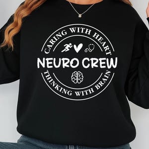 May include: Black sweatshirt with a white circular design. The design includes the words "NEURO CREW" and the phrases "CARING WITH HEART" and "THINKING WITH BRAIN". The design also features a running figure, a heart, a stethoscope, and a brain.