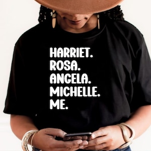 May include: Black t-shirt with white text: HARRIET. ROSA. ANGELA. MICHELLE. ME. The shirt is a crew neck style.