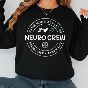 Pode incluir: Camisola preta com um gráfico circular branco que diz "Brain Mode: Always On, Neuro Crew, Neuro Life, Every Day". O gráfico inclui uma figura a correr, um coração, um estetoscópio e uma ilustração do cérebro.