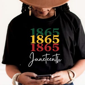May include: Black t-shirt with the word "Juneteenth" in a cursive font. The shirt features the numbers "1865" stacked in green, yellow, and red, representing the colours of the Pan-African flag.