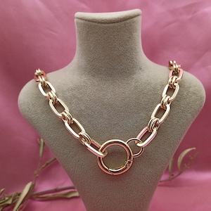 Könnte beinhalten: Eine goldfarbene Halskette mit großen ovalen Gliedern und einem kreisförmigen Verschluss. Die Halskette wird auf einer grauen Büste vor einem rosa Hintergrund präsentiert. Die Kette hat eine polierte Oberfläche, die das Licht reflektiert.
