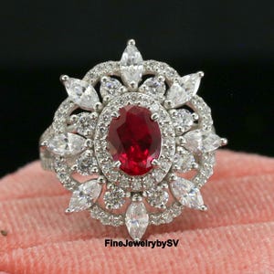 May include: A silver ring with a large oval ruby center stone, surrounded by smaller white gemstones in a floral design. The ring is displayed on a pink velvet ring box. The text "FineJewelrybySV" is visible.