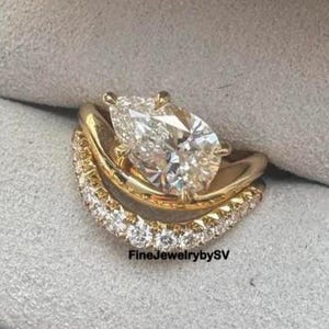May include: A gold engagement ring set with a pear-shaped diamond and a curved band with small diamonds. The rings are displayed in a blue velvet ring box with a gray interior. The text "FineJewelrybySV" is visible.