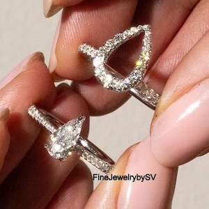 Peut inclure: Deux bagues en diamant, chacune avec une pierre centrale en forme de poire. L'une a un diamant en forme de poire massif, l'autre un halo de petits diamants en forme de poire. Les deux ont des bandes serties de diamants. Les bagues sont tenues par une personne.