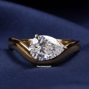 Puede incluir: Un anillo de oro con un diamante en forma de pera. El diamante está engastado en una banda de oro que se curva alrededor de la piedra. El anillo se muestra sobre una superficie azul. El texto "FineJewelrybySV" es visible.