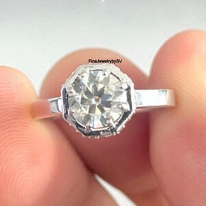 May include: A close-up of a silver ring with a large, clear, faceted gemstone. The gemstone is set in an octagonal bezel. The ring is held between fingers. The text "FineJewelrybySV" is visible.