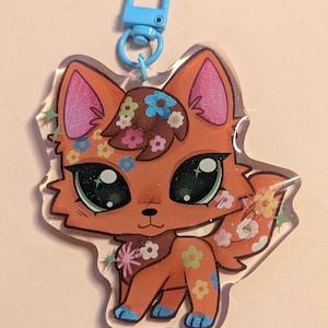 Könnte beinhalten: Ein klarer Acryl-Schlüsselanhänger mit einem Cartoon-Fuchs mit orangefarbenem Fell und floralen Akzenten. Der Fuchs hat große grüne Augen, rosa Ohren und blaue Füße. Oben ist ein blauer Verschluss angebracht. Der Schlüsselanhänger ist ein skurriles Accessoire.