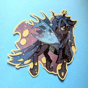 May include: A holographic sticker featuring a dark-colored, winged equine character with a crown, flowing mane, and tattered wing design. The illustration incorporates shades of purple, blue, and black with a sparkling star-shaped overlay effect.