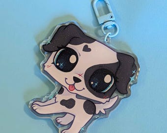 May include: A clear acrylic keychain featuring a cartoon dog with black and white spots. The dog has large, sparkling eyes, a pink tongue, and a small heart detail. The keychain has a light blue clasp.