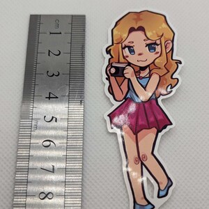 May include: A sticker of a cartoon woman with blonde hair, wearing a blue and pink dress, holding a camera. She has blue eyes and a pink blush on her cheeks.