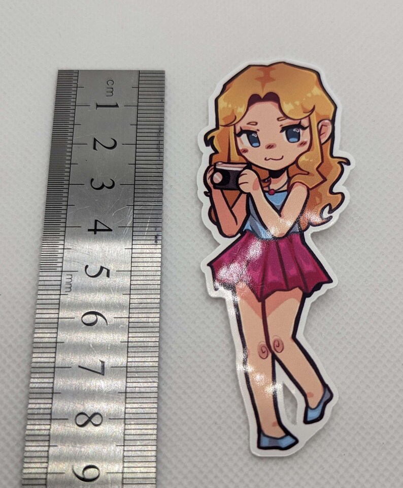 May include: A sticker of a cartoon woman with blonde hair, wearing a blue and pink dress, holding a camera. She has blue eyes and a pink blush on her cheeks.