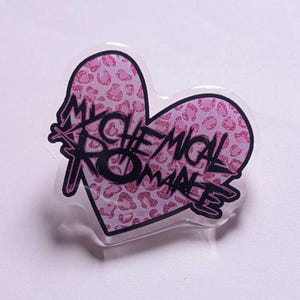 May include: A heart-shaped pin with a pink leopard print design. The pin features the text "My Chemical Romance" in black, stylized lettering. The pin is made of clear plastic.