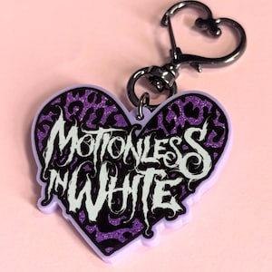 Pode incluir: Um chaveiro em forma de coração com as palavras "Motionless In White" em letras brancas estilizadas. O coração tem um design de estampa de leopardo roxo com detalhes de glitter e uma borda roxa. O chaveiro tem um fecho prateado.