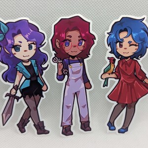 May include: Three cartoon character stickers with different colored hair and outfits. The character on the left has purple hair and is wearing a blue jacket and black pants. The character in the middle has red hair and is wearing a blue jumpsuit. The character on the right has blue hair and is wearing a red dress.
