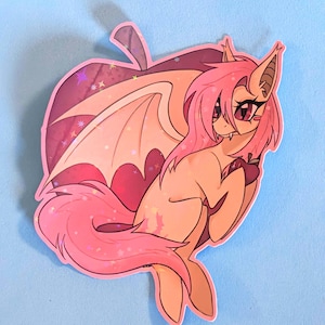 May include: A sticker featuring a cartoon creature with bat-like ears and wings, a pink mane, and a long tail. The creature is set against a pink apple-shaped background with star accents. The sticker has a glittery finish.