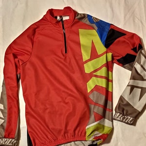 Könnte beinhalten: Rotes Langarm-Radtrikot mit geometrischem Design in Grau, Schwarz und Gelb. Das Trikot hat einen Viertelreißverschluss.