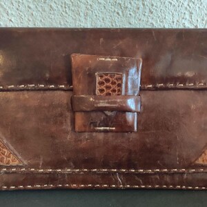 Può includere: Una pochette in pelle marrone di forma rettangolare. La pochette presenta una chiusura con patta strutturata e cuciture decorative. Accenti triangolari di un materiale marrone più chiaro sono visibili sul davanti.