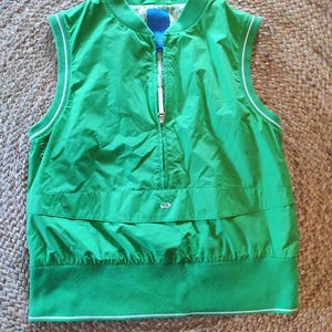 Può includere: Un gilet verde senza maniche con zip frontale, finiture bianche e collo alto. Il gilet ha un orlo inferiore in pile e un accento blu all'interno del colletto. Il gilet è realizzato in materiale leggero.