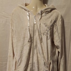 Könnte beinhalten: Ein cremefarbener Velours-Kapuzenpullover mit einem Blumendesign auf der linken Seite. Der Hoodie hat einen weißen Reißverschluss und weiße Kordeln.