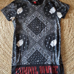 Könnte beinhalten: Schwarzes T-Shirt mit einem Bandana-Muster in Weiß und Schwarz. Das Shirt hat einen seitlichen Reißverschluss und den roten Text "CRIMINVL DVMMGE" am Saum. Es hat kurze Ärmel und einen Rundhalsausschnitt. Das Design ist auffällig.