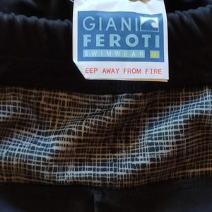 Può includere: Pantaloncini da bagno neri con un motivo geometrico e un'etichetta con la scritta "GIANI FEROTI SWIMWEAR XXL". L'etichetta include anche l'avvertenza "KEEP AWAY FROM FIRE".