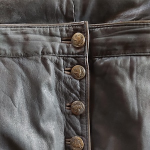 Könnte beinhalten: Nahaufnahme einer dunkelbraunen Lederhose mit Knopfleiste. Die Knöpfe sind aus Metall und haben ein dekoratives Design.