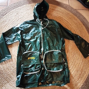 Könnte beinhalten: Dunkelgrüne Regenjacke mit Kapuze, weißem Reißverschluss und zwei Vordertaschen. Auf einer der Taschen ist das Wort "STIHL" in Gelb aufgedruckt. Die Jacke besteht aus einem glänzenden, wasserdichten Material.