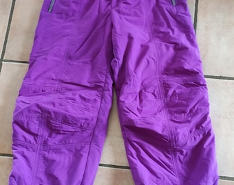 May include: Vibrant purple ski trousers with multiple pockets and zipped closures. The trousers feature an elasticated waistband and elasticated cuffs at the ankles. The fabric has a quilted appearance. These ski trousers are designed for winter sports and outdoor activities.
