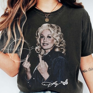 May include: A charcoal gray short-sleeve t-shirt featuring a graphic print of a blonde woman with voluminous hair wearing a denim jacket. The graphic shows the woman gesturing with both middle fingers. The text Dolly is printed below.