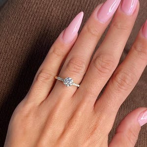 May include: A close-up of a hand wearing a gold ring with a large, round diamond. The hand is also wearing a gold watch. The nails are painted a light pink color and are long and pointed.