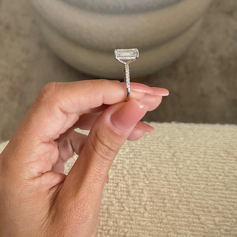 Op de afbeelding: Een zilveren ring met een grote, rechthoekige, heldere edelsteen. De ring heeft een dunne band met kleine, heldere edelstenen die in de band zijn gezet.