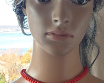 Könnte beinhalten: Ein leuchtend rotes Choker-Halsband. Die Halskette besteht aus einem geflochtenen Material, das ein strukturiertes Aussehen erzeugt. Das Choker-Halsband liegt eng am Hals an und zeigt seine kräftige Farbe und sein schlichtes Design. Der Hintergrund ist unscharf, wodurch der Fokus auf dem Schmuck liegt.