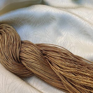 Puede incluir: Primer plano de un haz de cordón dorado, bien enrollado y retorcido. El cordón está sobre una tela color crema con un patrón sutil y repetitivo. El cordón parece estar hecho de muchos hilos finos.