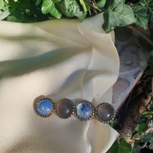 Pode incluir: Uma presilha de cabelo dourada com quatro pedras redondas e iridescentes. As pedras estão emolduradas em um quadro decorativo e repousam sobre um tecido creme. A folhagem verde envolve a presilha, criando um fundo natural.