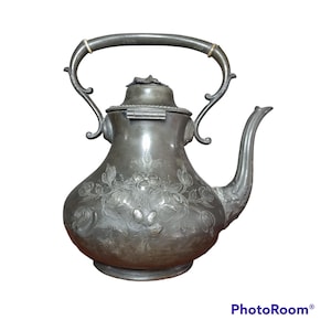 Op de afbeelding: Een zilveren theepot met een bloemenmotief en een scharnierend deksel. De theepot heeft een gebogen tuit en een handvat met een decoratief scrollontwerp.