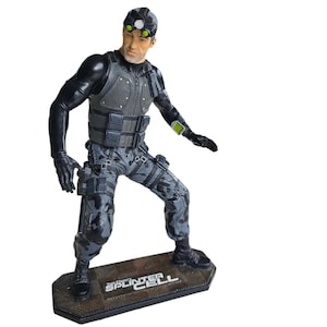 Op de afbeelding: Een zwart en grijs actiefiguur van Sam Fisher uit de Tom Clancy's Splinter Cell videogame-serie. Het figuur is in een hurkende positie met een pistool in zijn rechterhand. Het figuur staat op een zwarte basis met de woorden "Tom Clancy's Splinter Cell" erop gedrukt.