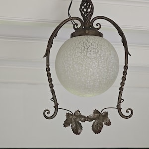 Op de afbeelding: Oorspronkelijke hanglamp in antieke stijl. Voorzien van een matglazen bol, ondersteund door een smeedijzeren frame met decoratieve blad- en wijnstokaccenten. De armatuur heeft een gedraaide metalen bovenkant en gebogen armen.