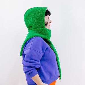 Könnte beinhalten: Ein grüner Kapuzen-Schal mit einem lilafarbenen Sweatshirt. Der Schal ist aus einem weichen, flauschigen Material.