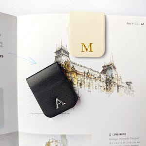 Peut inclure: Deux marque-pages en cuir. L'un est noir avec la lettre "A" argentée, l'autre est crème avec la lettre "M" dorée. Les marque-pages sont sur une page de livre avec un dessin d'un bâtiment.