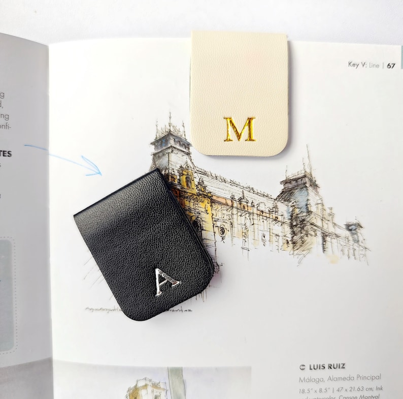 May include: Two leather book page holders. One is black with a silver letter "A", the other is cream-colored with a gold letter "M". The page holders are on a book page with a drawing of a building.