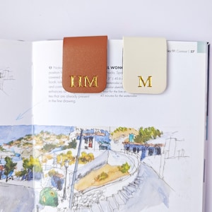 May include: Two leather book page markers, one tan and one cream, are clipped onto a book. The tan marker has the gold letters "HM", and the cream marker has the gold letter "M". The book is open to a watercolor illustration.