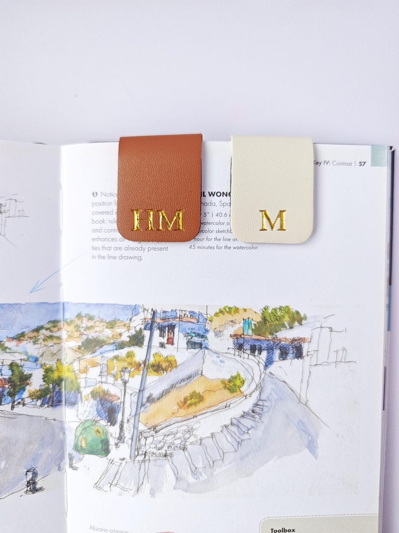 May include: Two leather book page markers, one tan and one cream, are clipped onto a book. The tan marker has the gold letters "HM", and the cream marker has the gold letter "M". The book is open to a watercolor illustration.
