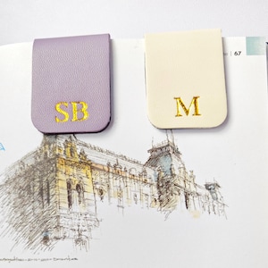 May include: Two leather magnetic bookmarks. One is lavender with gold embroidered initials "SB", the other is cream with gold embroidered initial "M". The bookmarks are on a book with a sketch of a building.