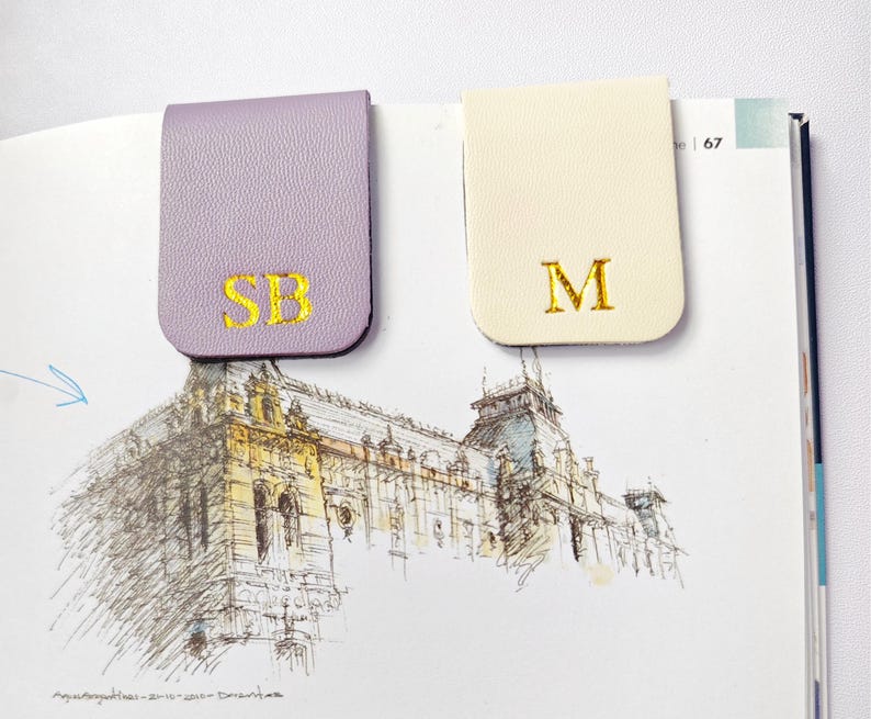 May include: Two leather magnetic bookmarks. One is lavender with gold embroidered initials "SB", the other is cream with gold embroidered initial "M". The bookmarks are on a book with a sketch of a building.