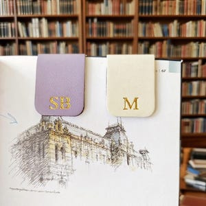 Puede incluir: Dos marcadores magnéticos de cuero. Uno es lavanda con las iniciales doradas bordadas "SB", el otro es crema con la inicial dorada bordada "M". Los marcadores están en un libro con un boceto de un edificio.